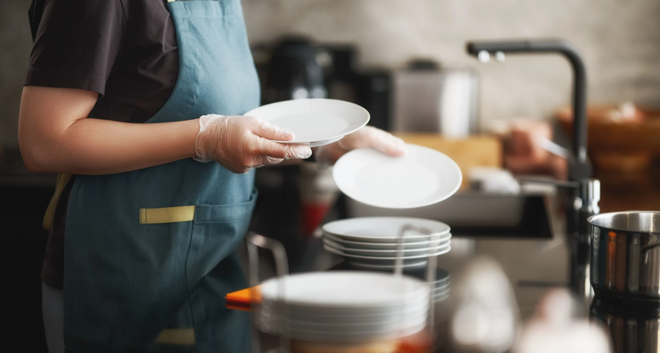 Restaurant employee wearing gloves and apron while handling clean dishes in a commercial kitchen